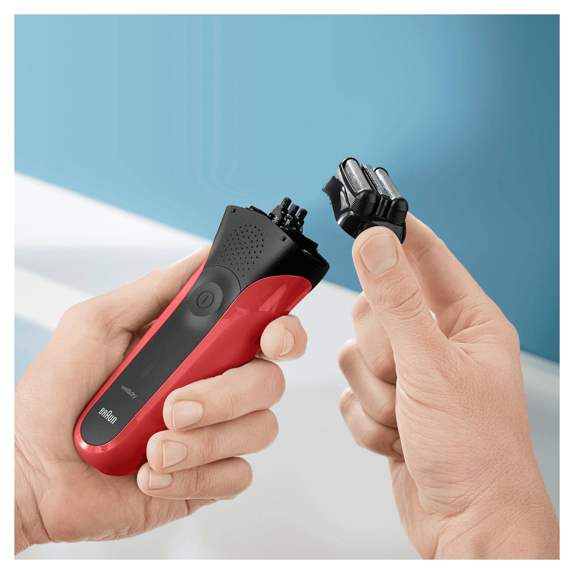 Eine Hand, die einen roten und schwarzen elektrischen Rasierer vor einem hellblauen Hintergrund hält, während eine andere Hand einen abnehmbaren Rasierkopf darüber präsentiert.
