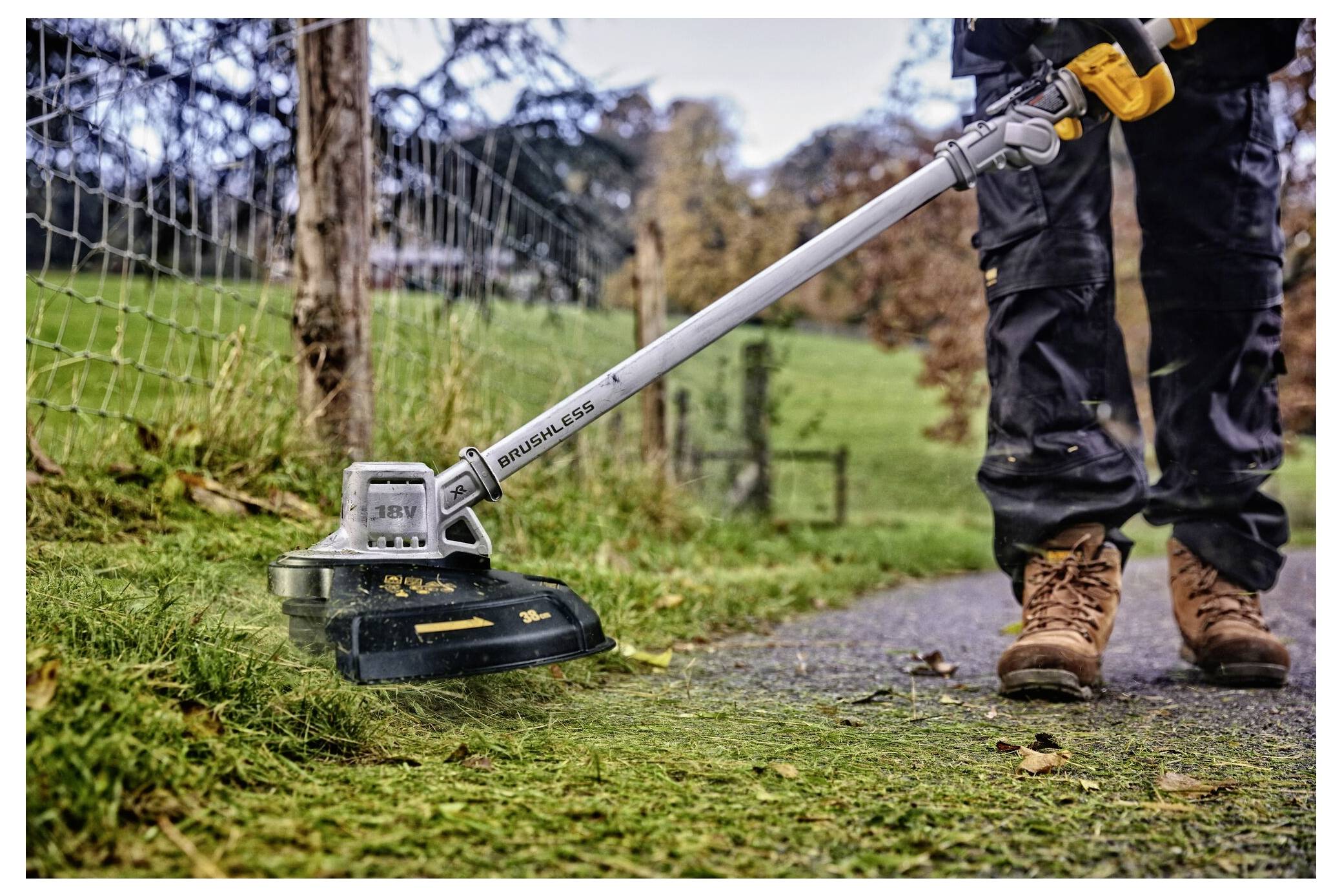 Eine Person verwendet eine Motorsense, um Gras entlang eines Weges in einem Park zu schneiden. Sie trägt Arbeitshose und Stiefel, mit Herbstbäumen im Hintergrund.