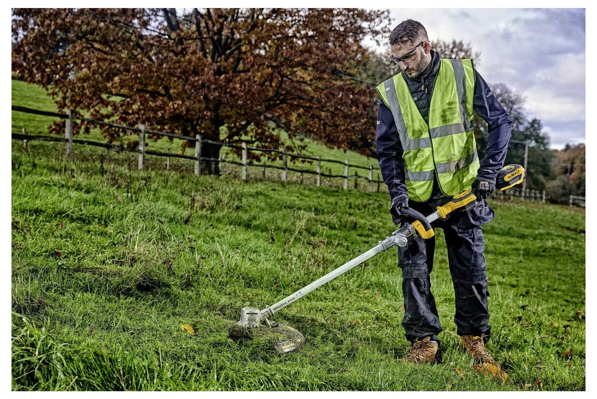 Eine Person in Sicherheitsausrüstung benutzt einen Freischneider an einem grasigen Hang, mit einem Holzzaun und Herbstbäumen im Hintergrund.