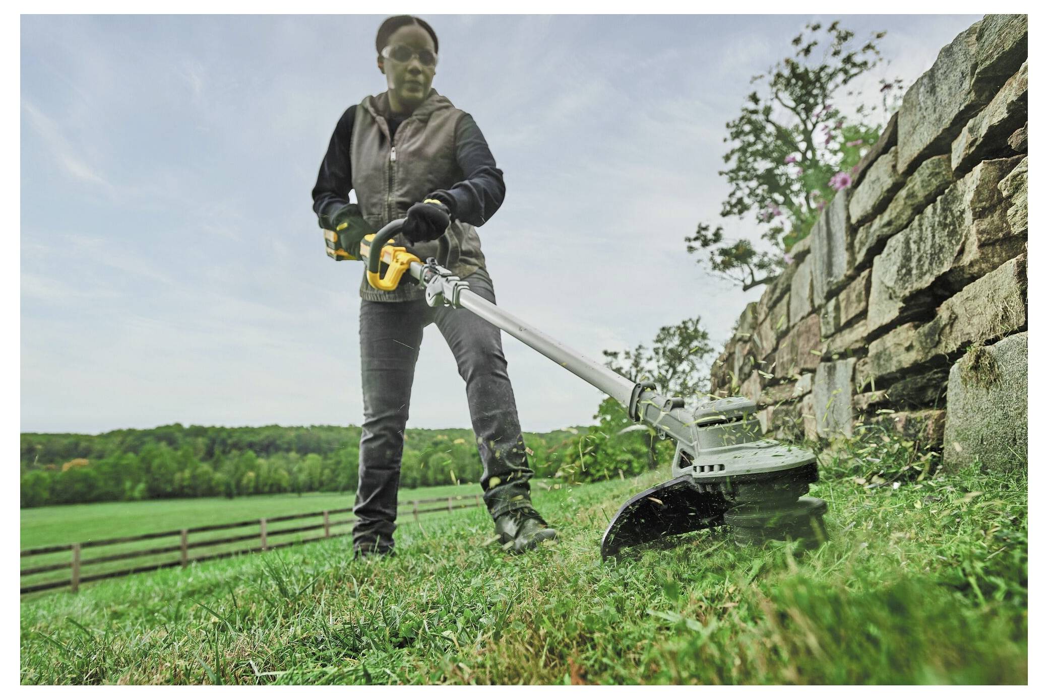 Eine Person trimmt Gras mit einem Rasentrimmer in der Nähe einer Steinmauer, vor einer Kulisse aus einem grünen Feld und Bäumen unter einem bewölkten Himmel.