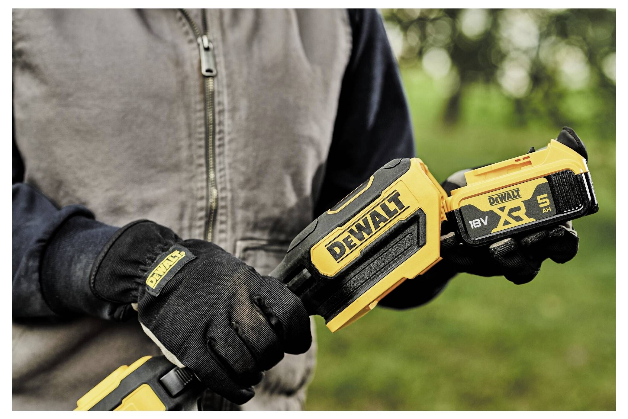 Eine Person in einer Jacke und Handschuhen hält ein gelbes DeWalt-Elektrowerkzeug mit der Beschriftung „18V XR 5 Ah" im Freien, wobei Bäume im Hintergrund zu sehen sind.