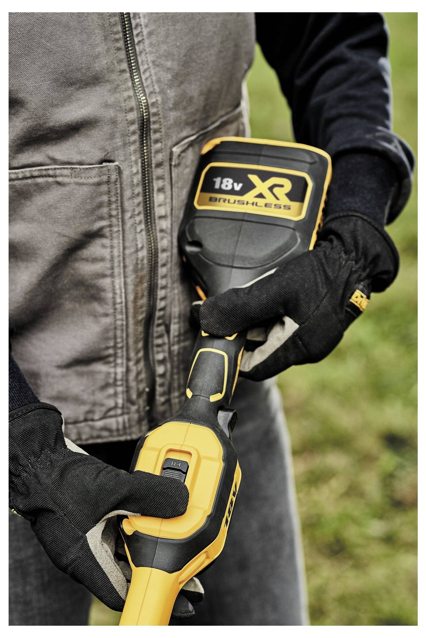 Eine Person trägt Arbeitshandschuhe und hält ein gelb-schwarzes 18-V-Akku-Elektrowerkzeug im Freien, mit Gras im Hintergrund.