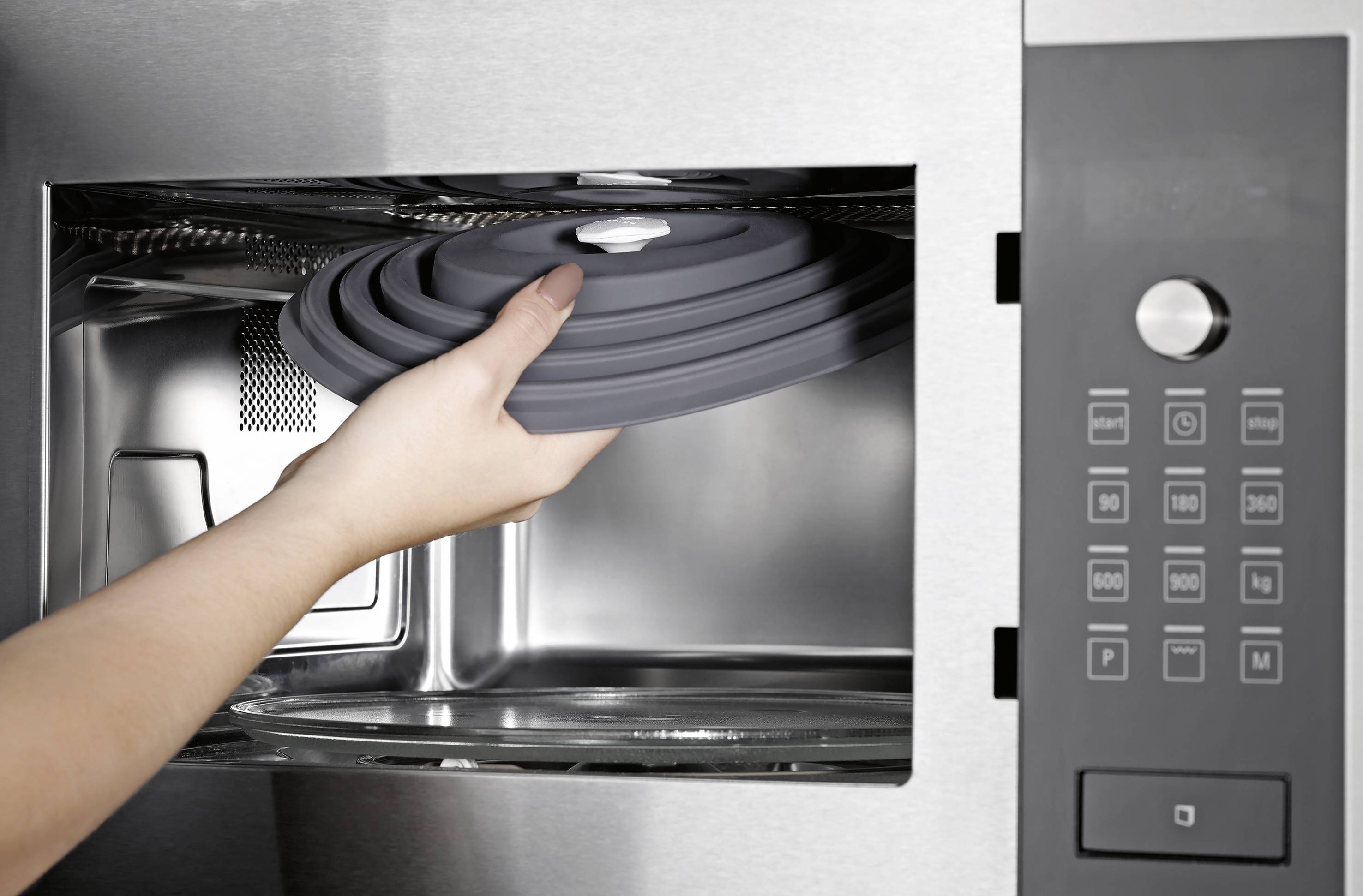 Eine Person schiebt einen Silikondeckel in einen Edelstahl-Mikrowellenherd und bereitet sich darauf vor, ein Gericht zum Kochen oder Aufwärmen abzudecken.