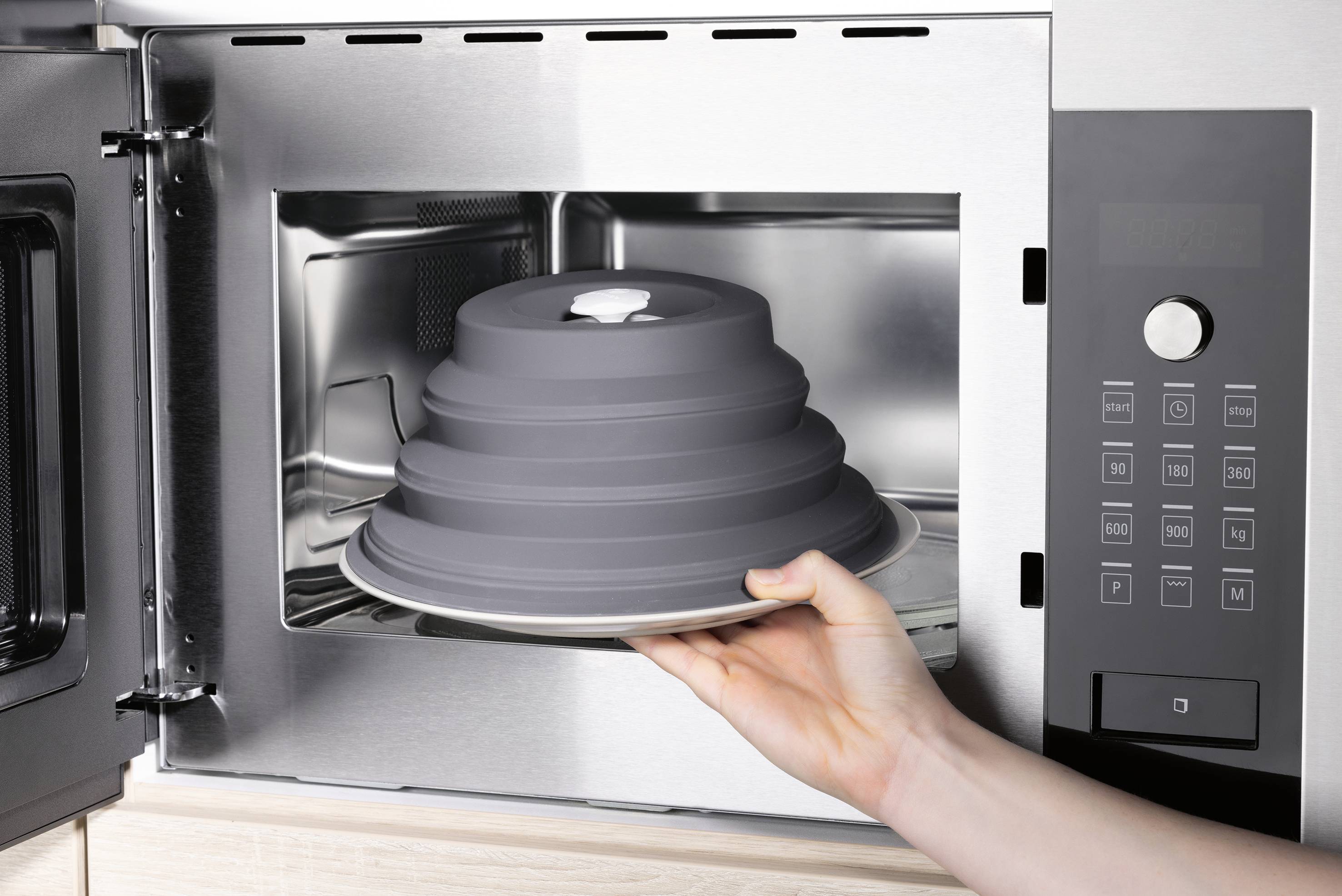 Eine Hand, die einen stapelbaren Silikonlebensmitteldeckel in einen Edelstahl-Mikrowellenherd legt und damit sichere Lebensmittelaufbewahrung und Aufwärmnutzung demonstriert.