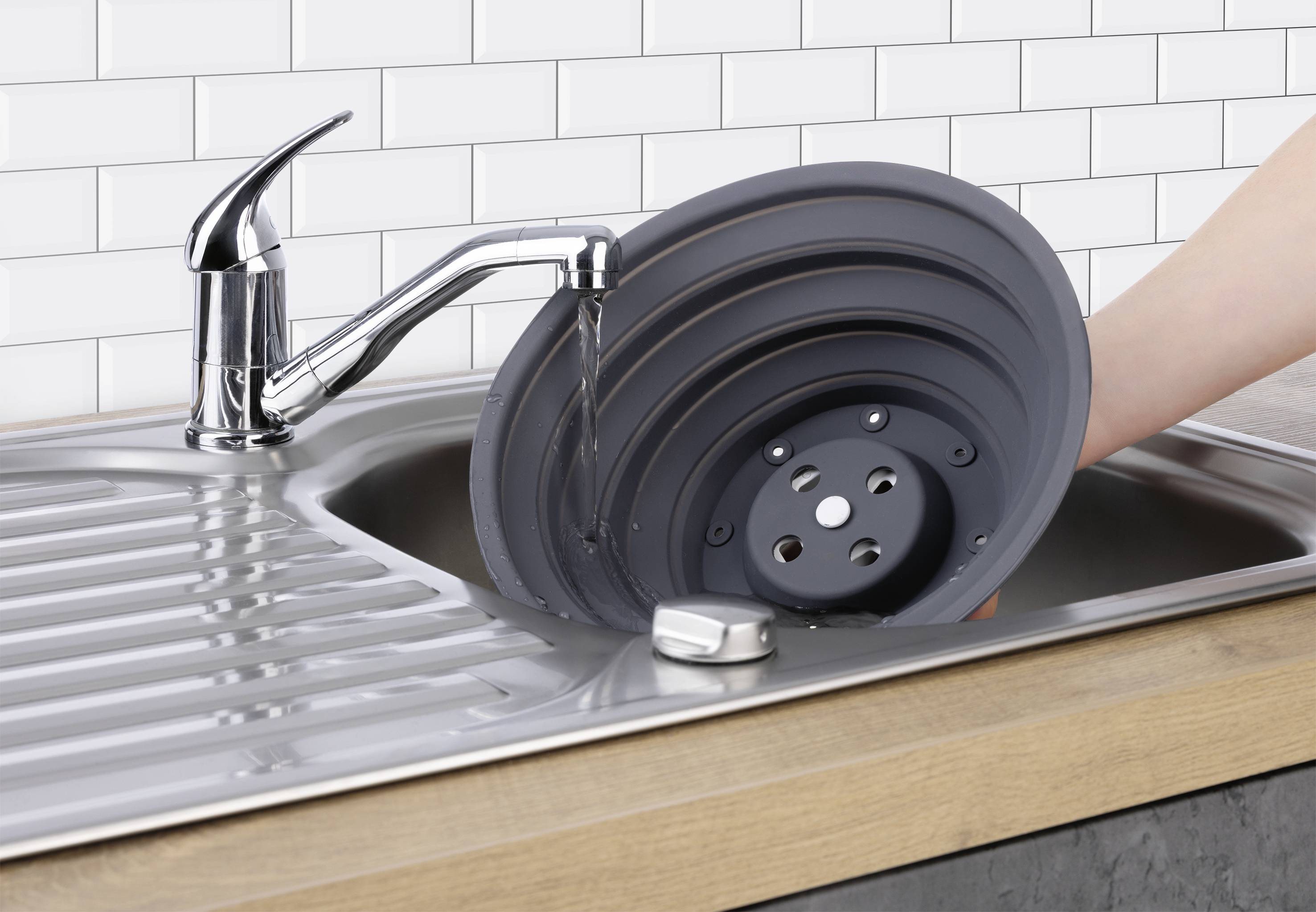 Eine Hand wäscht ein zusammenklappbares, rundes schwarzes Abtropfgestell unter einem Küchenhahn mit fließendem Wasser, über einem Edelstahlspülbecken.