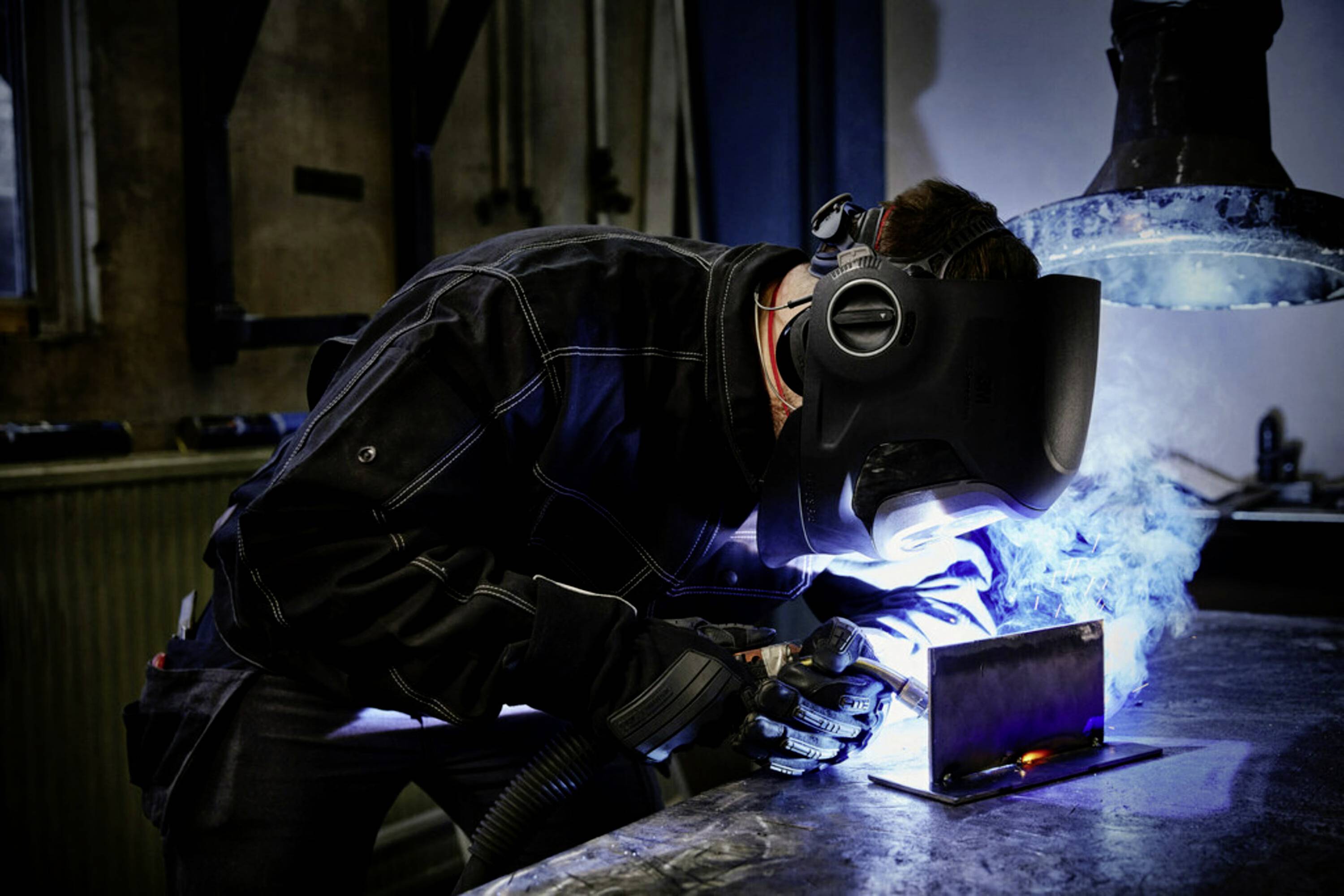 Eine Person in Schutzausrüstung schweißt Metall auf einer Werkbank, wobei helle Funken und Rauch sichtbar sind. Industrielle Werkstattumgebung.