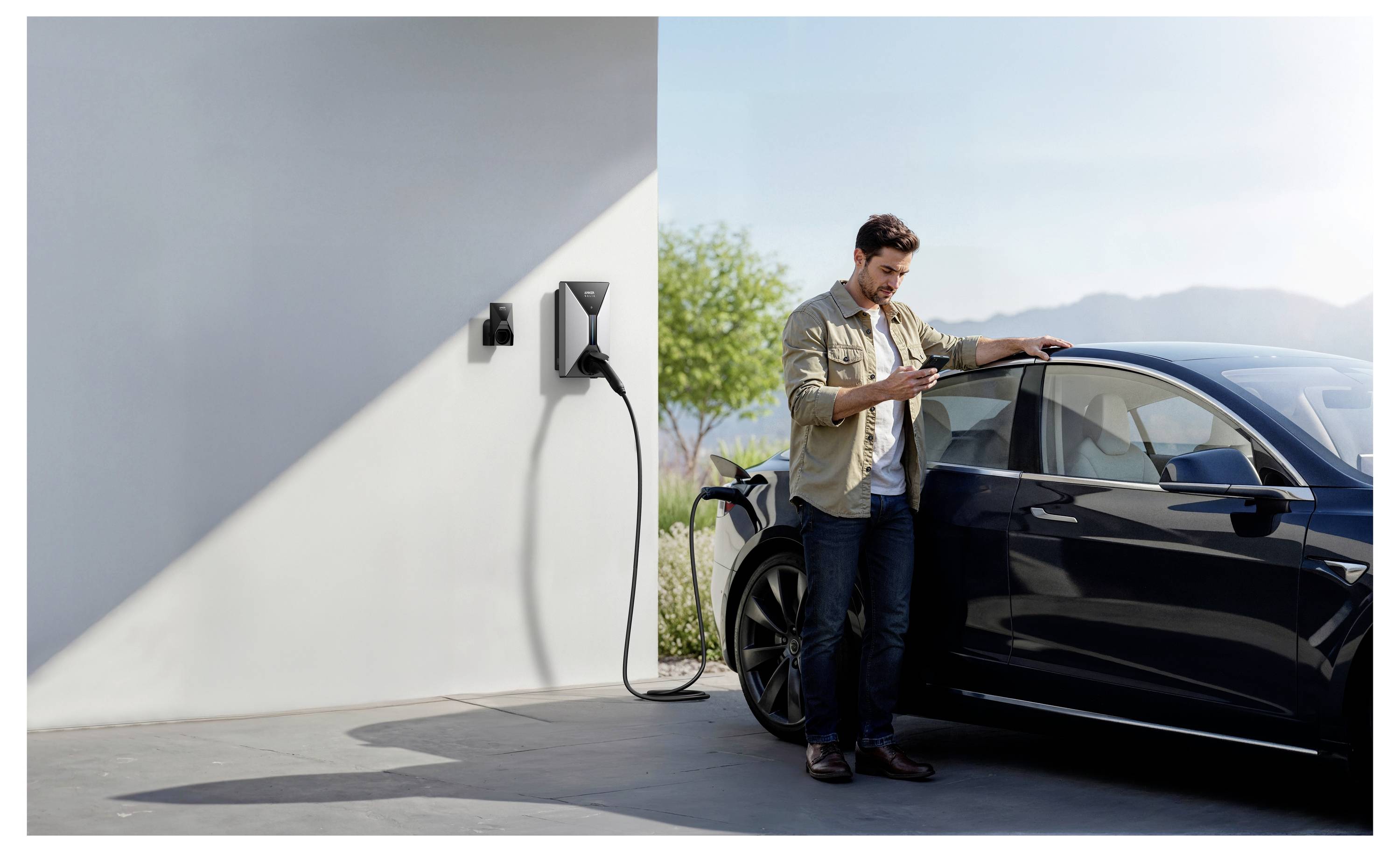 Ein Mann steht neben einem geparkten Elektroauto, lehnt sich dagegen und schaut auf sein Telefon. Das Auto ist an einer wandmontierten Ladestation angeschlossen.
