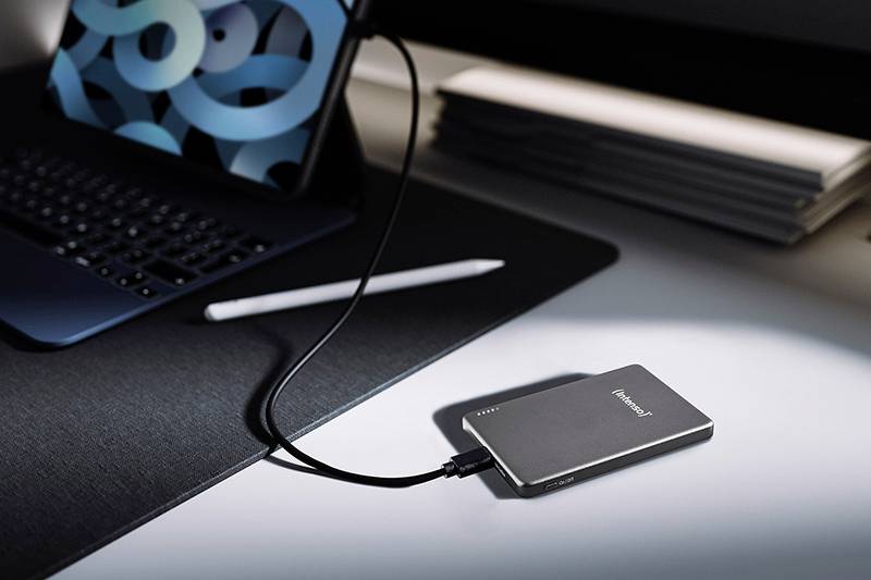 Eine schlanke externe Festplatte, die an einem Laptop angeschlossen ist, mit einem Griffel in der Nähe auf einem Schreibtisch. Der Laptop zeigt einen abstrakten gemusterten Hintergrund.