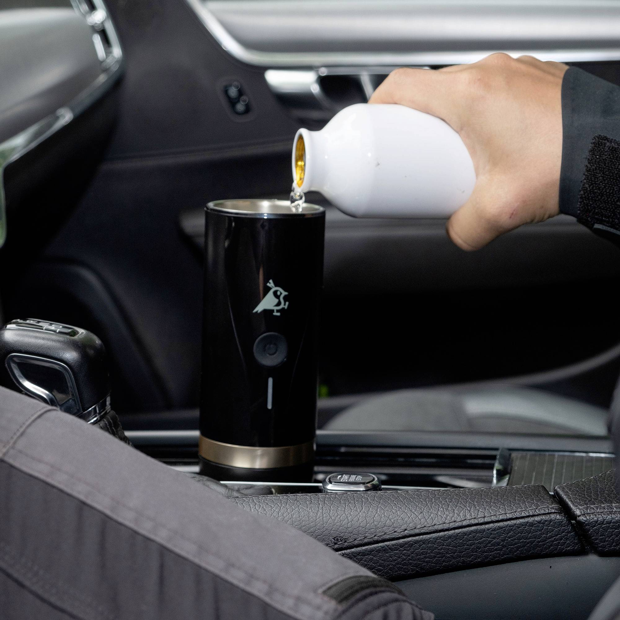 Eine Hand gießt Flüssigkeit aus einer weißen Flasche in einen tragbaren schwarzen Heizgerät im Inneren eines Autos, was die Vorbereitung eines Getränks andeutet.