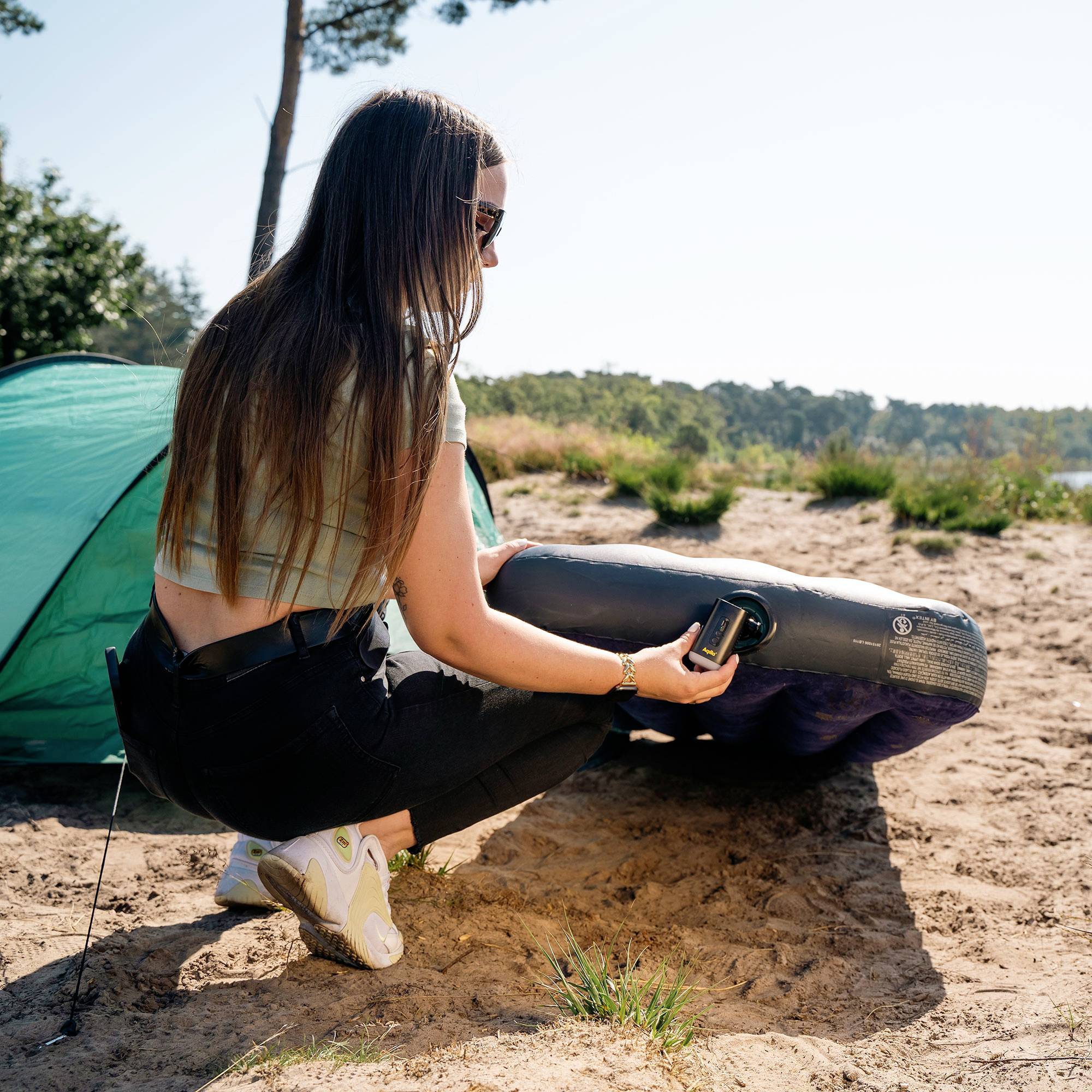 Eine Person, die eine Luftmatratze mit einer Pumpe in der Nähe eines grünen Zeltes an einem sonnigen Tag aufpumpt, umgeben von Bäumen und Gras.