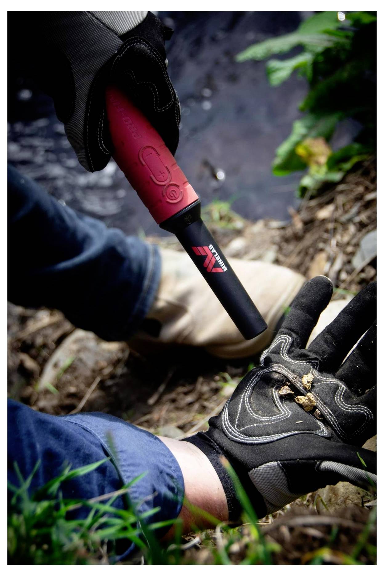 Person mit Handschuhen, die einen tragbaren Metallsuchgerät nahe dem Boden benutzt, wobei Laub und Wasser im Hintergrund sichtbar sind.