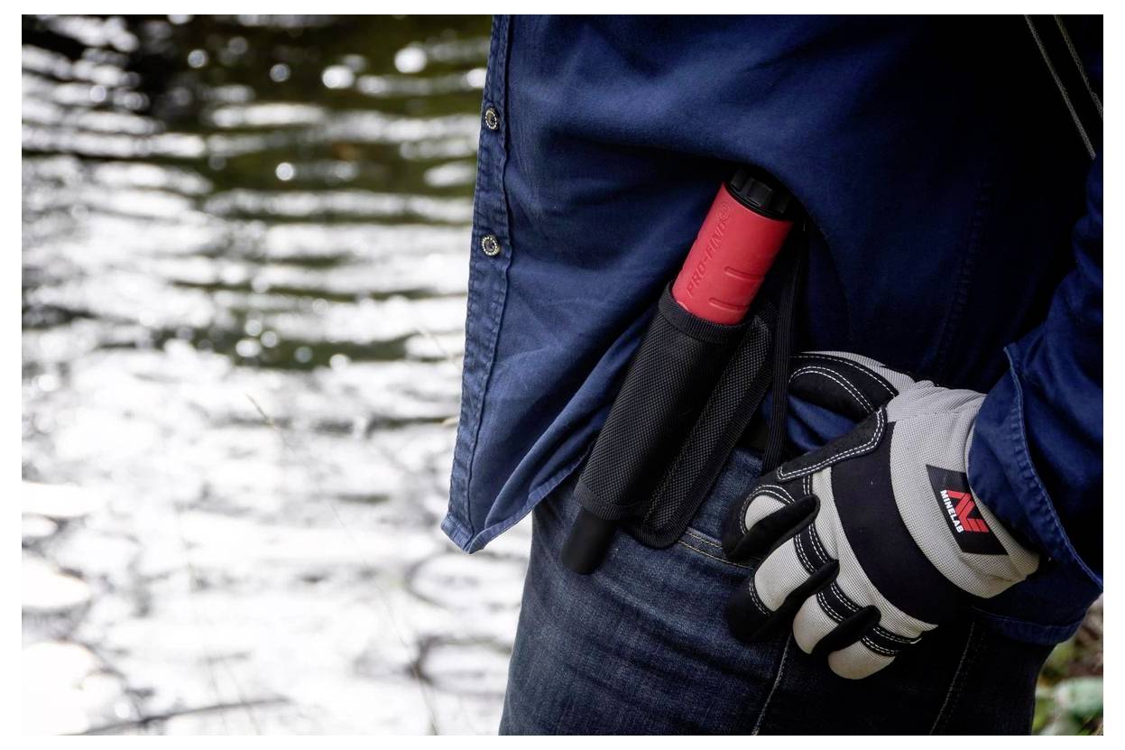 Eine Person in einem blauen Hemd und Handschuhen steht neben einem Gewässer, mit einer roten und schwarzen Scheide an ihrem Gürtel.