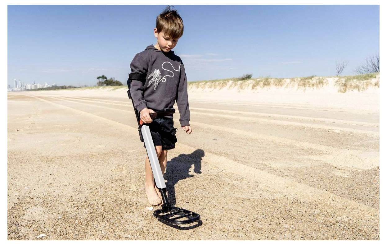 Ein kleiner Junge geht an einem sandigen Strand und hält einen Metalldetektor, während er aufmerksam den Boden auf einem klaren Tag absucht.