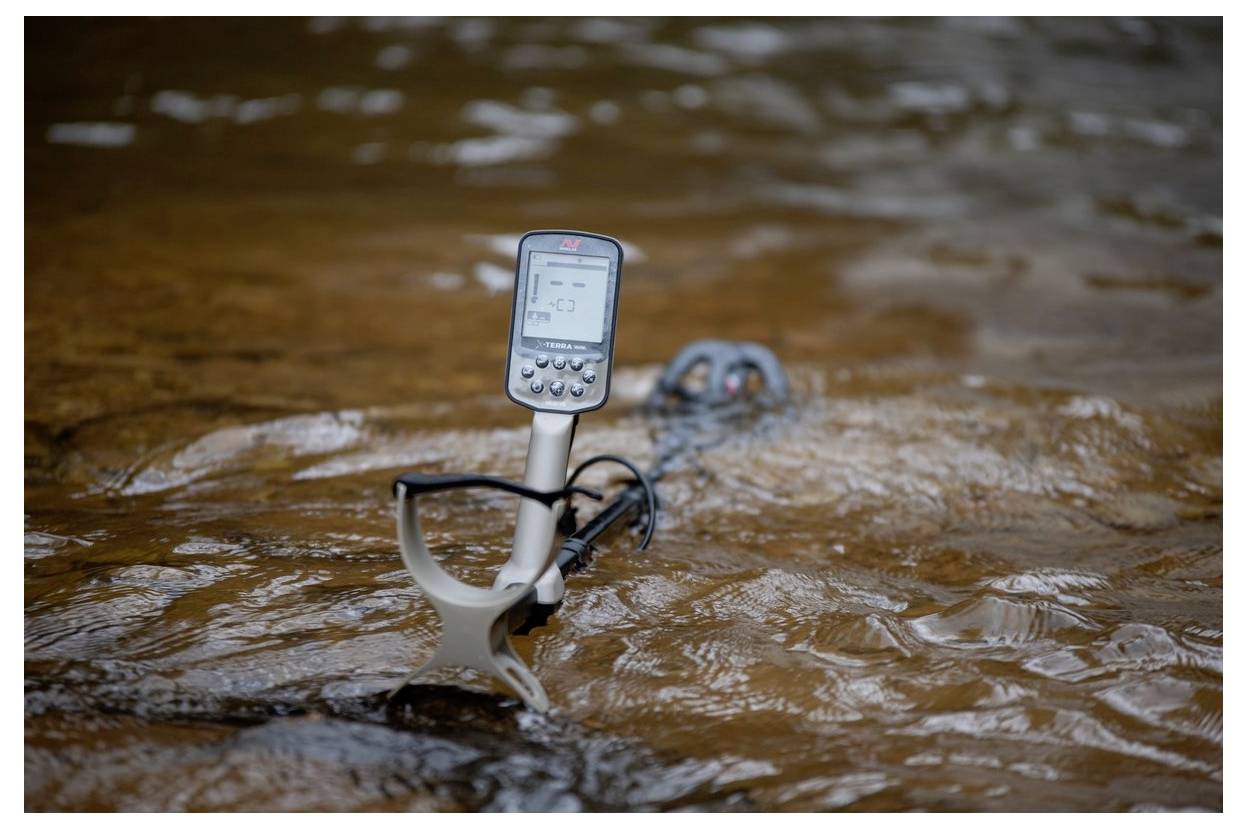Ein Metallsuchgerät, das teilweise im Wasser versenkt ist und dessen Verwendung in einer seichten Fluss- oder Bachlandschaft anzeigt.