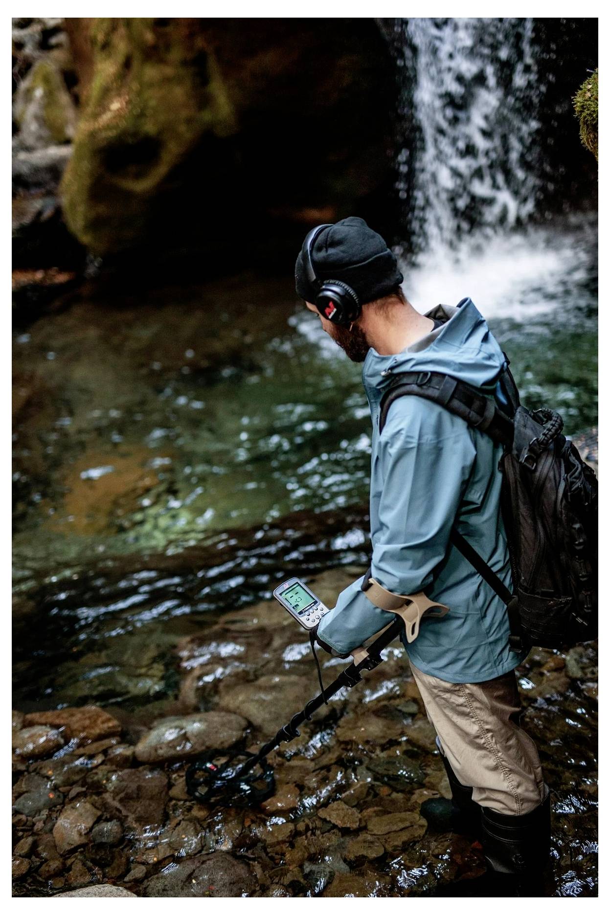 Eine Person, die einen Metalldetektor in einem felsigen Bach unter einem Wasserfall benutzt, trägt eine blaue Jacke, einen Rucksack und Kopfhörer.