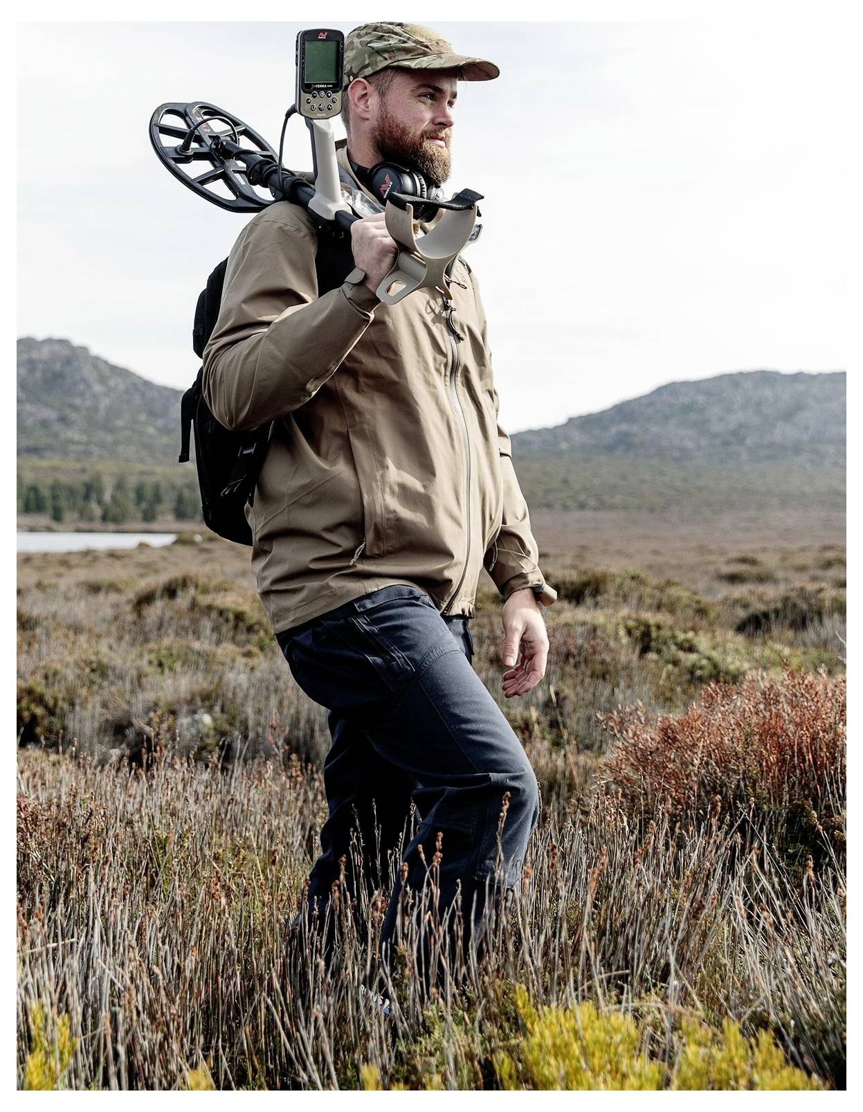 Eine Person mit Hut und Rucksack steht auf einem grasigen Feld und hält einen Metalldetektor, während im Hintergrund Hügel und ein bewölkter Himmel zu sehen sind.