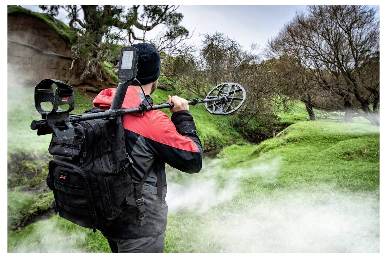 Eine Person mit einem Rucksack und einem Metalldetektor steht in einer nebeligen, grasigen Landschaft und konzentriert sich darauf, nach Metallgegenständen im Boden zu suchen.