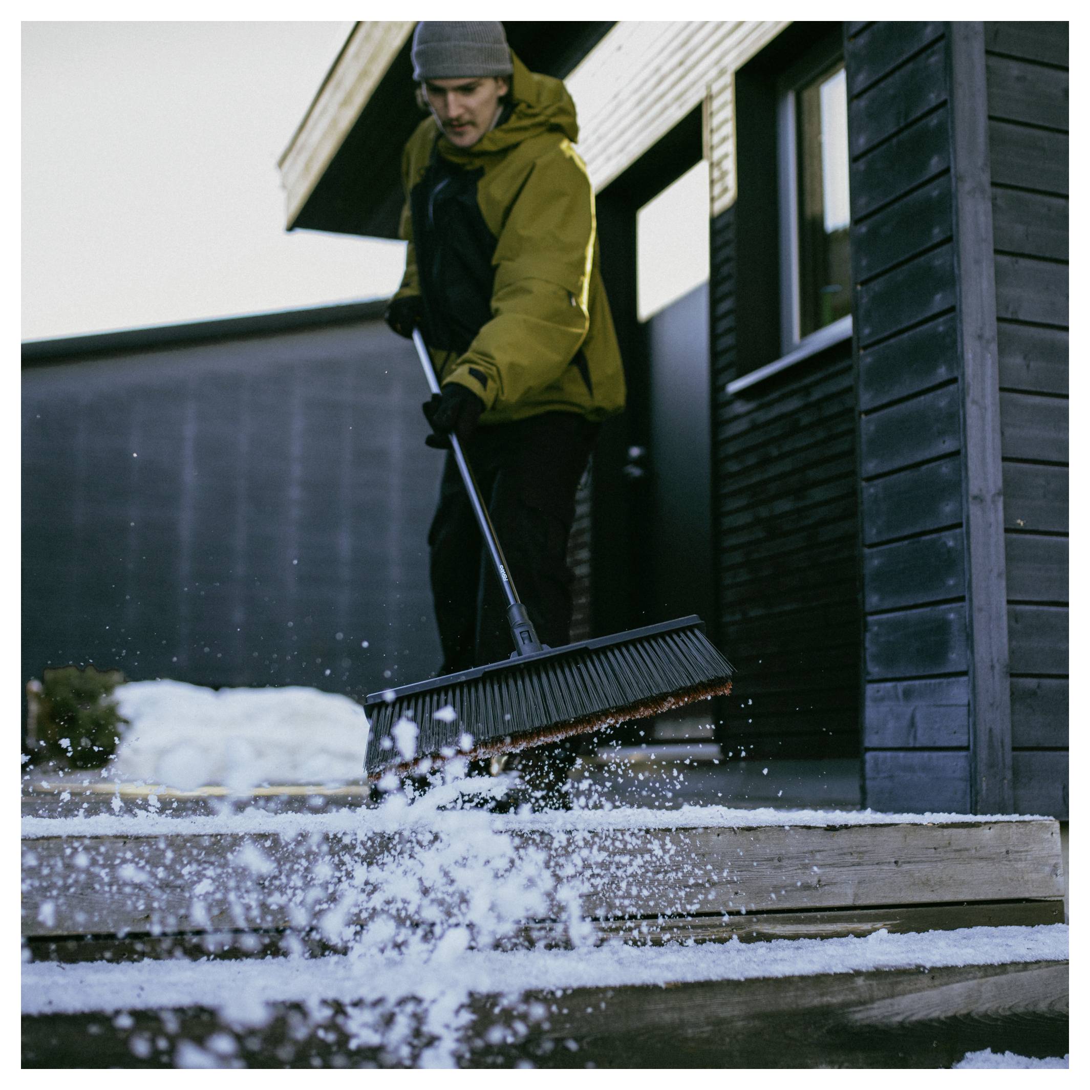 Eine Person in Winterkleidung, die Schnee von einer Holzterrasse vor einem modernen Gebäude schaufelt.