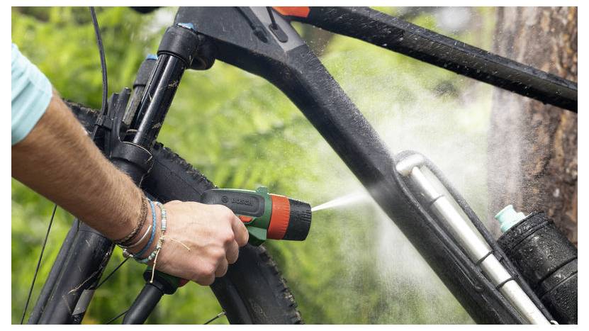 Eine Person reinigt ein schwarzes Fahrrad mit einer Sprühdüse und entfernt Schmutz und Ablagerungen. Der Hintergrund zeigt Grünpflanzen und deutet auf eine Outdoor-Umgebung hin.