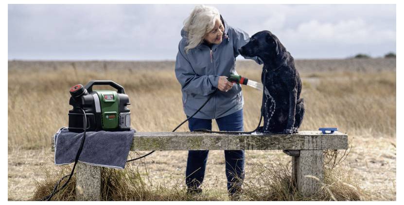 Eine Frau wäscht einen schwarzen Hund auf einer Steinbank in einem grasbewachsenen Feld mit einer tragbaren Waschmaschine. Ein Handtuch und ein Reinigungsgerät befinden sich neben ihr.
