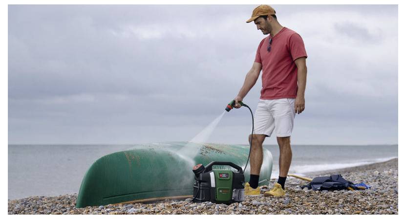 Eine Person in einem roten Hemd und einer roten Kappe wäscht ein grünes Kanu mit einem tragbaren Hochdruckreiniger an einem felsigen Strand, mit dem Meer im Hintergrund.