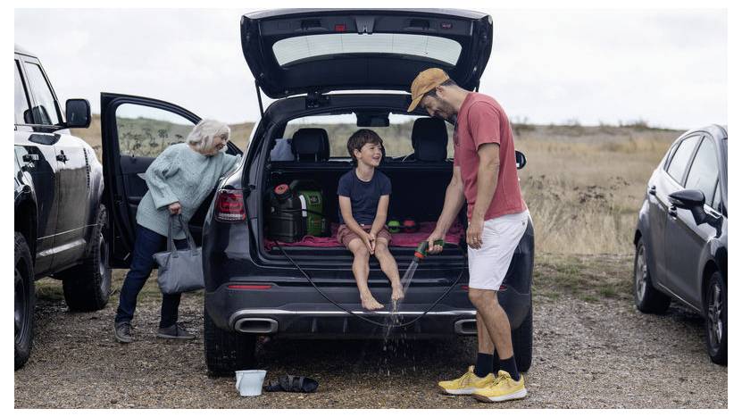 Ein Mann spült die Füße eines Kindes mit Wasser aus einem Schlauch am Heck eines geparkten SUVs ab. Eine ältere Frau steht in der Nähe und beobachtet die Szene. Outdoor-Umgebung.