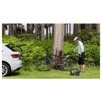 Eine Person in Radfahrkleidung verwendet einen tragbaren Hochdruckreiniger, um ein Fahrrad zu reinigen, das an einen Baum in einem bewaldeten Gebiet gelehnt ist.