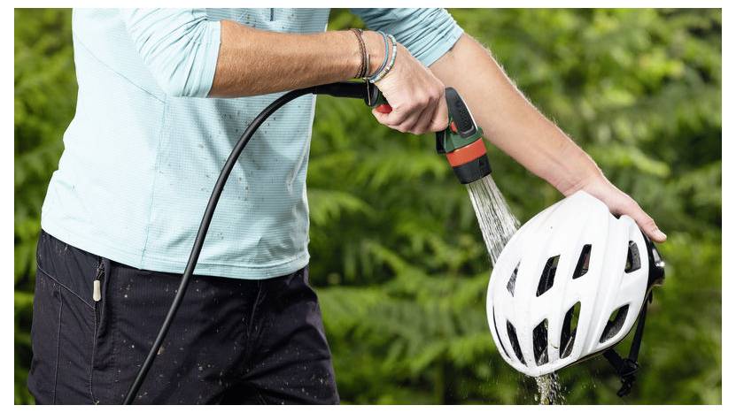 Eine Person, die einen weißen Fahrradhelm mit einem Gartenschlauch im Freien abspült, umgeben von grünem Laub.
