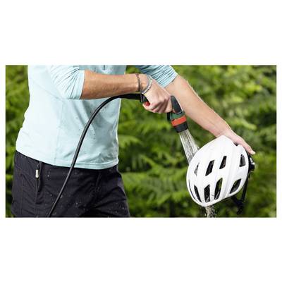 Eine Person, die einen weißen Fahrradhelm mit einem Gartenschlauch im Freien abspült, umgeben von grünem Laub.