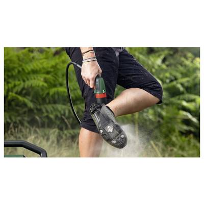 Eine Person benutzt einen Hochdruckreiniger, um Schmutz von einem Schuh zu entfernen, während sie im Freien steht, umgeben von Grünpflanzen.