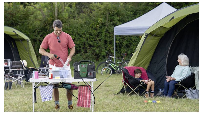 Ein Mann bereitet Essen an einem Tisch mit Campingausrüstung zu. Eine Frau und ein Kind sitzen auf Klappstühlen in der Nähe von Zelten auf einer Rasenfläche.