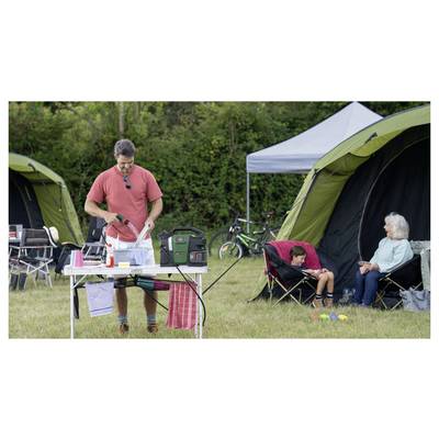 Ein Mann bereitet Essen an einem Tisch mit Campingausrüstung zu. Eine Frau und ein Kind sitzen auf Klappstühlen in der Nähe von Zelten auf einer Rasenfläche.