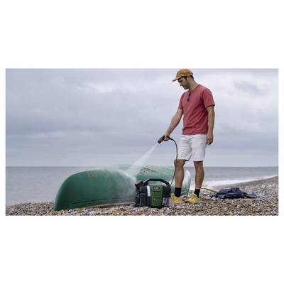 Ein Mann in einem roten T-Shirt und kurzen Hosen benutzt einen tragbaren Hochdruckreiniger, um ein grünes Kajak an einem Kieselstrand zu reinigen.