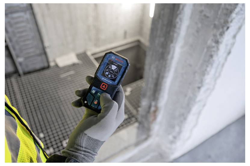 Eine Person hält einen Laser-Entfernungsmesser auf einer Baustelle. Das Gerät zeigt Messdaten an. Der Hintergrund zeigt Betonwände.