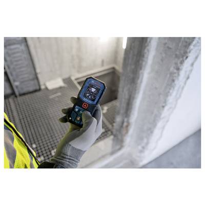Eine Person hält einen Laser-Entfernungsmesser auf einer Baustelle. Das Gerät zeigt Messdaten an. Der Hintergrund zeigt Betonwände.