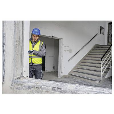 Ein Bauarbeiter in einer Sicherheitsweste und mit Schutzhelm untersucht ein Gerät in einem teilweise renovierten Gebäude neben einer Treppe.
