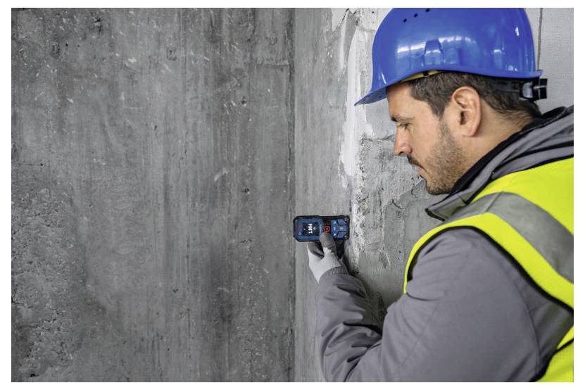 Eine Person mit blauem Helm und gelber Sicherheitsweste verwendet ein Gerät, um eine Betonwand zu vermessen oder zu inspizieren.