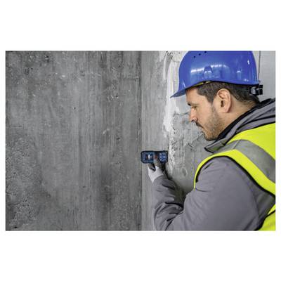 Eine Person mit blauem Helm und gelber Sicherheitsweste verwendet ein Gerät, um eine Betonwand zu vermessen oder zu inspizieren.