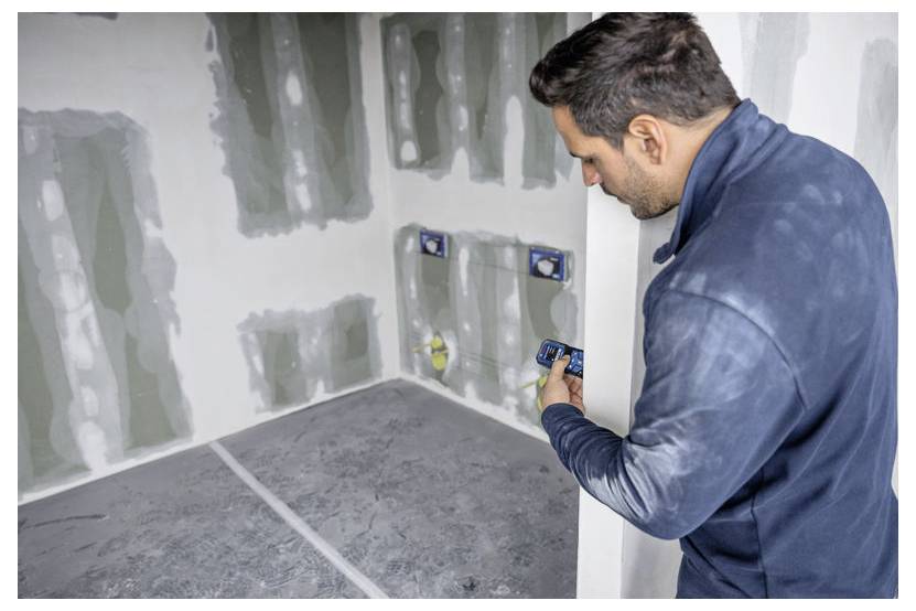 Eine Person, die ein Gerät hält und einen teilweise im Bau oder in der Renovierung befindlichen Raum mit unfertig wirkenden Wänden und sichtbaren Elektrodosen inspiziert.