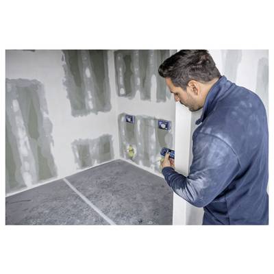 Eine Person, die ein Gerät hält und einen teilweise im Bau oder in der Renovierung befindlichen Raum mit unfertig wirkenden Wänden und sichtbaren Elektrodosen inspiziert.