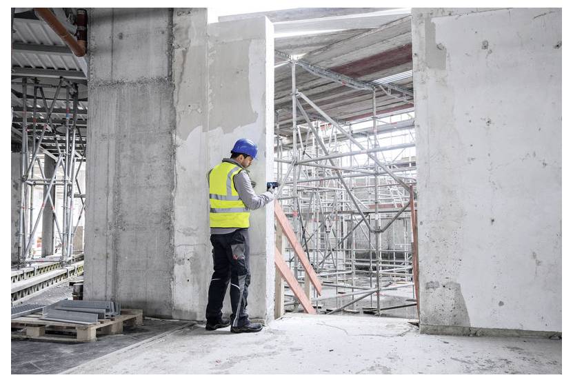 Ein Bauarbeiter mit blauem Helm und gelber Warnweste inspiziert eine Baustelle mit Gerüsten und Betonwänden in einem industriellen Umfeld.