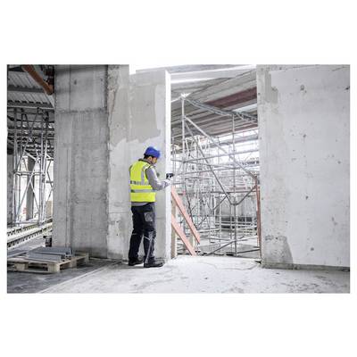Ein Bauarbeiter mit blauem Helm und gelber Warnweste inspiziert eine Baustelle mit Gerüsten und Betonwänden in einem industriellen Umfeld.