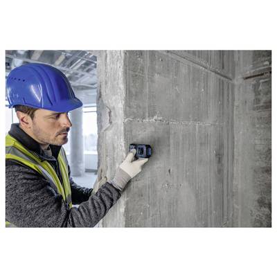 Ein Bauarbeiter mit blauem Schutzhelm und Sicherheitsweste verwendet ein Messwerkzeug an einer Betonwand in einem sich im Bau befindlichen Gebäude.