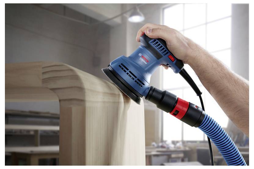 Eine Hand benutzt einen blauen Exzenterschleifer, um die Kante einer Holzoberfläche in einer Werkstatt zu glätten. Der Staub wird über einen angeschlossenen Schlauch aufgefangen.