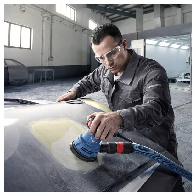 Ein Mann mit Schutzbrille schleift eine Motorhaube in einer Werkstatt und verwendet dabei einen elektrischen Schleifer. Der Raum hat einen modernen, industriellen Stil.
