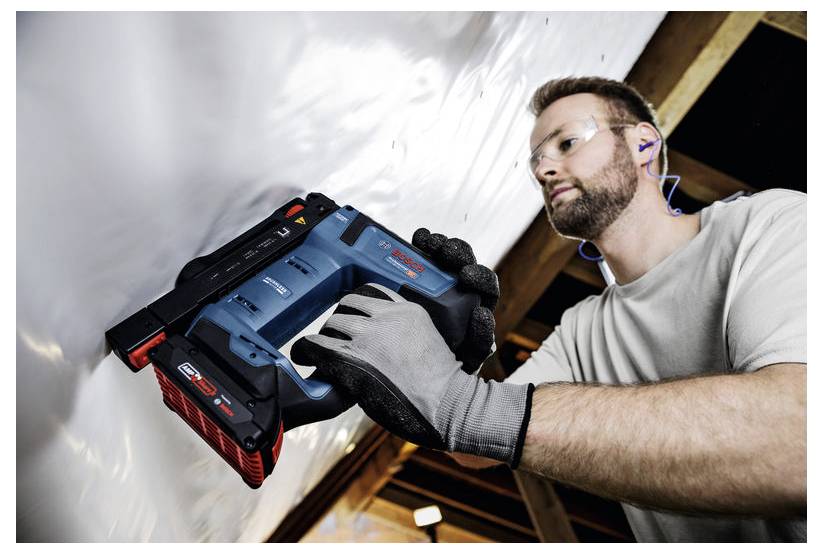 Eine Person mit Handschuhen und Sicherheitsausrüstung verwendet einen blauen, kabellosen elektrischen Nagler, um Material an der Decke einer Holzkonstruktion zu befestigen.