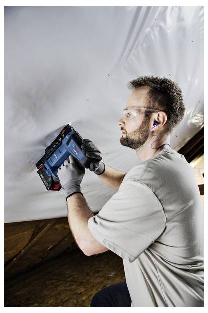 Eine Person mit Sicherheitsbrille und Handschuhen verwendet ein Elektrowerkzeug, um ein weißes Blatt an einer Oberfläche zu befestigen und konzentriert sich dabei aufmerksam auf die Aufgabe.