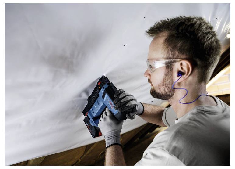 Eine Person mit Schutzbrille und Handschuhen verwendet eine blaue elektrische Tackerpistole, um ein weißes Material an einer Deckenstruktur zu befestigen.
