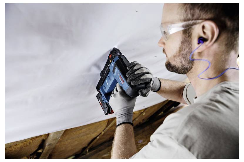 Eine Person mit Sicherheitsbrille und Gehörschutz verwendet ein Bosch-Werkzeug, um Material auf einer Holzoberfläche in einer Baustelle zu befestigen.