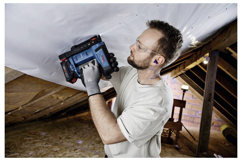 Eine Person mit Sicherheitsbrille und Handschuhen verwendet einen elektrischen Tacker, um ein weißes Blatt auf dem Dachboden an der Decke zu befestigen.